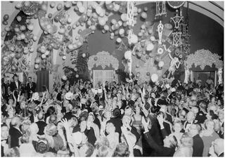 Black and white photo of a room full of people reaching up for hundreds of balloons