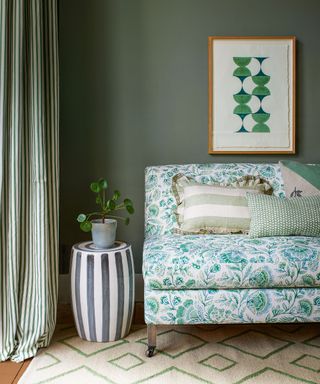 Green painted living room with green floral patterned sofa, striped side table, plant, cushions, artwork on wall