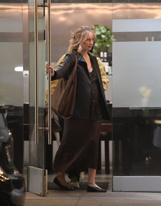 Jennifer Lawrence leaving dinner in Los Angeles with a friend wearing statement earrings, a black leather blazer, a brown skirt and top set, and black flats with a brown oversized bag.