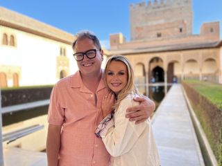 Alan Carr and Amanda Holden stand in the grounds of the Alhambra palace in Granada. Alan has his arm around Amanda's shoulder and she is leaning on his chest. They are both smiling warmly.