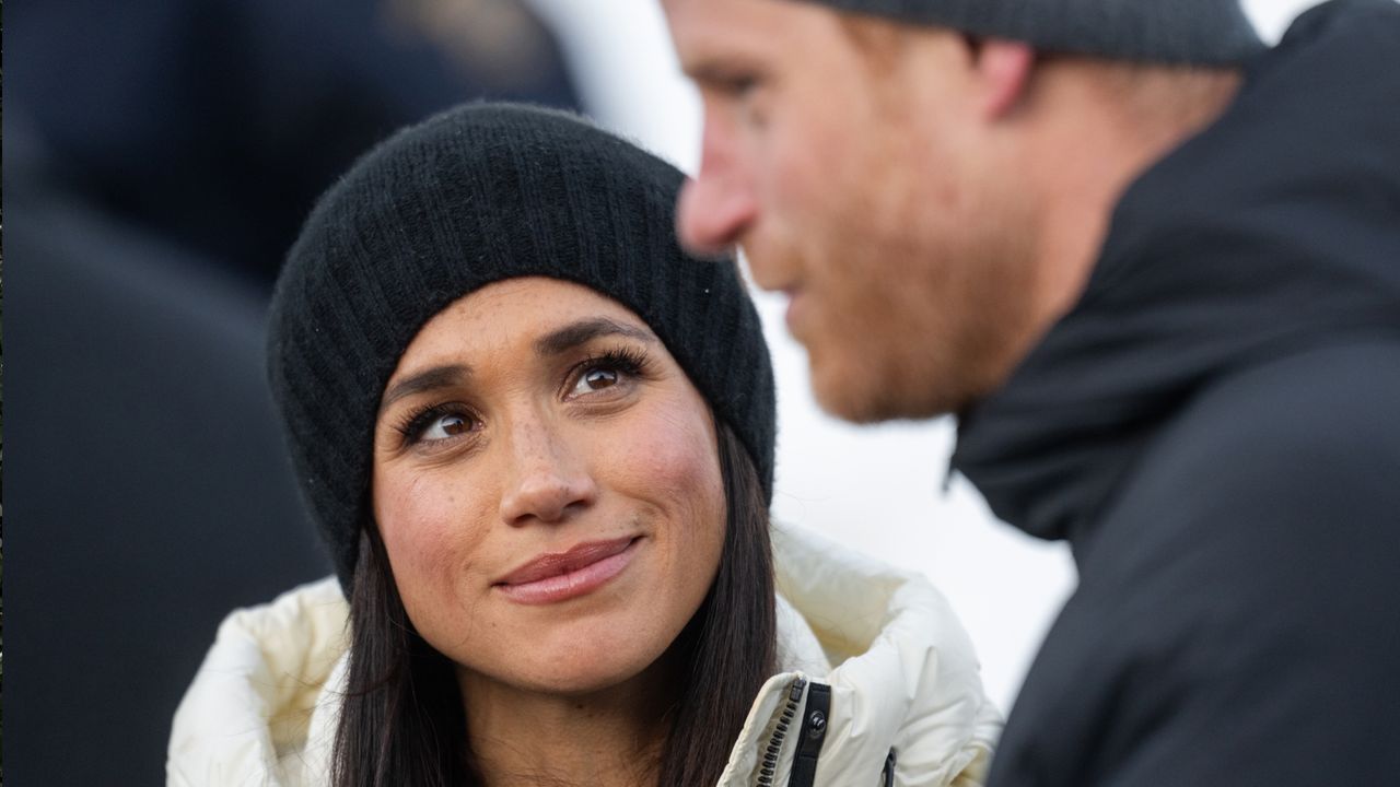 Meghan Markle wearing a black hat and white winter coat smiling up at Prince Harry, dressed in a black coat and hat