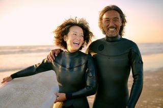 An older couple in black wetsuits smiles for the camera holding a surfboard on a beach at sunset.