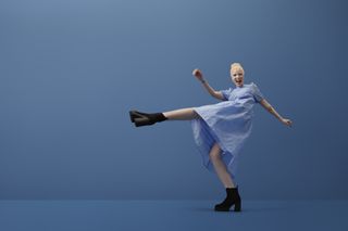 A woman wearing a pair of black, platform boots, kicking her leg out to the side in front of a blue background.