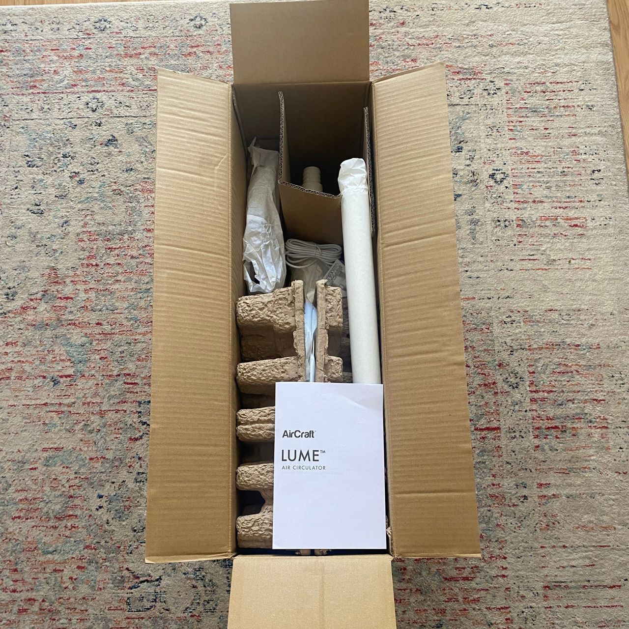 The opened packaging of the AirCraft LUME Quiet Air Circulator Fan on a beige, grey and red rug