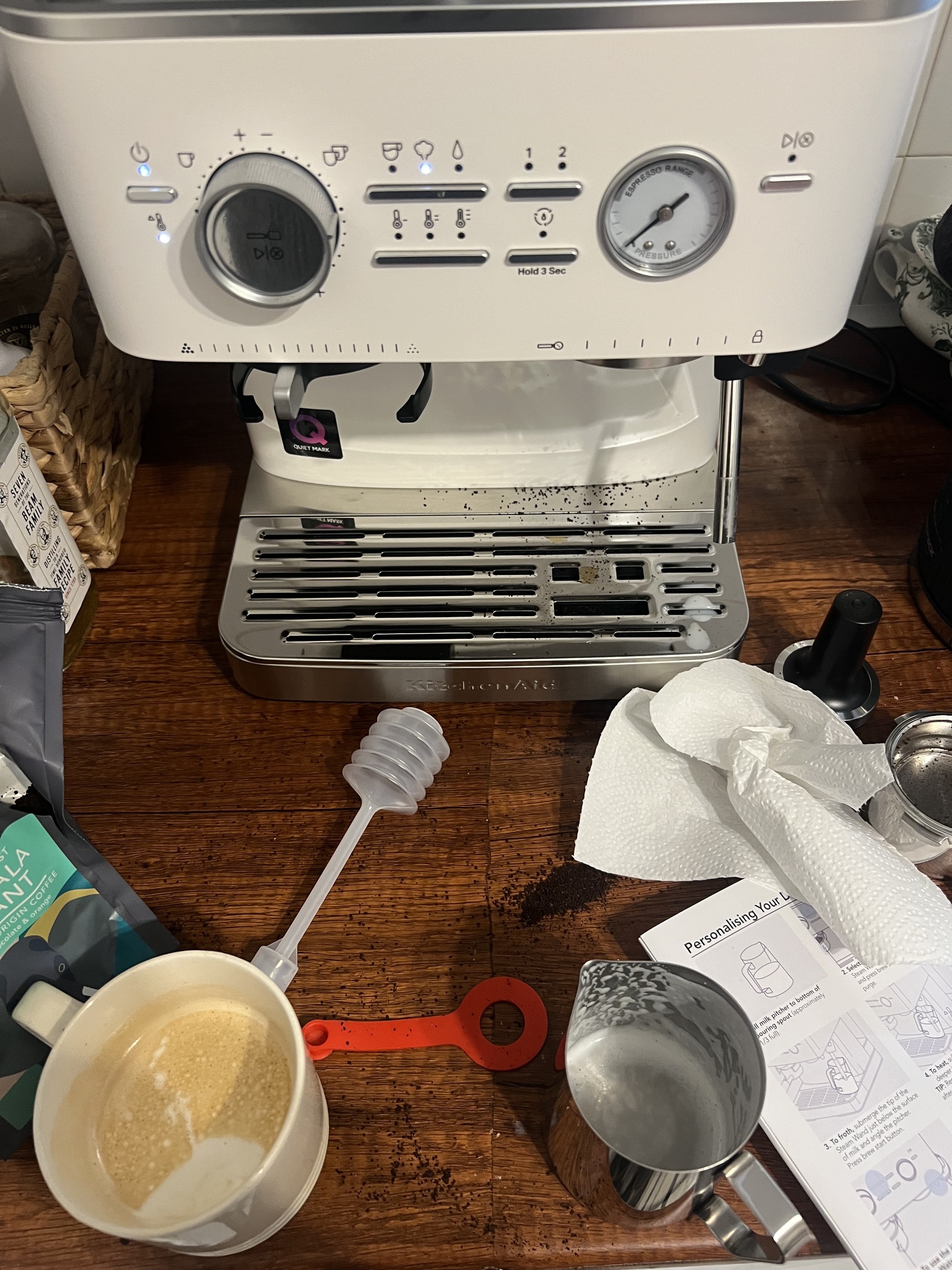 Image of a white coffee machine on a wooden countertop with coffee grinds spilled on the counter and a mess of coffee machine accessories around it.