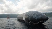 a giant black-tiled rocket lays afloat on the ocean with a boat and an island in the background.