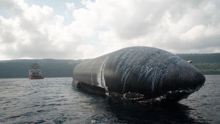 a giant black-tiled rocket lays afloat on the ocean with a boat and an island in the background.
