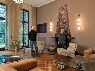 Tommaso Fantoni and Valeria Borsani standing in Villa Borsani’s living room - there is wood flooring, light coloured walls, tall doors with windows above, curtains, colourful flowers in a marble vase, wall lights, a fireplace, dark coloured wall art, glass tables and brown and cream seating
