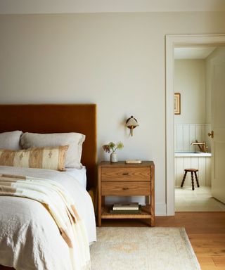 bedroom with cream walls and velvet headboard in warm brown tones and view to en suite bathroom with cream panelling