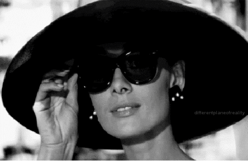 Audrey Hepburn pulls down her sunglasses to look at something.