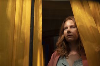 The Woman In The Window: Anna Fox (Amy Adams) stands in a darkened house, peering through a curtain