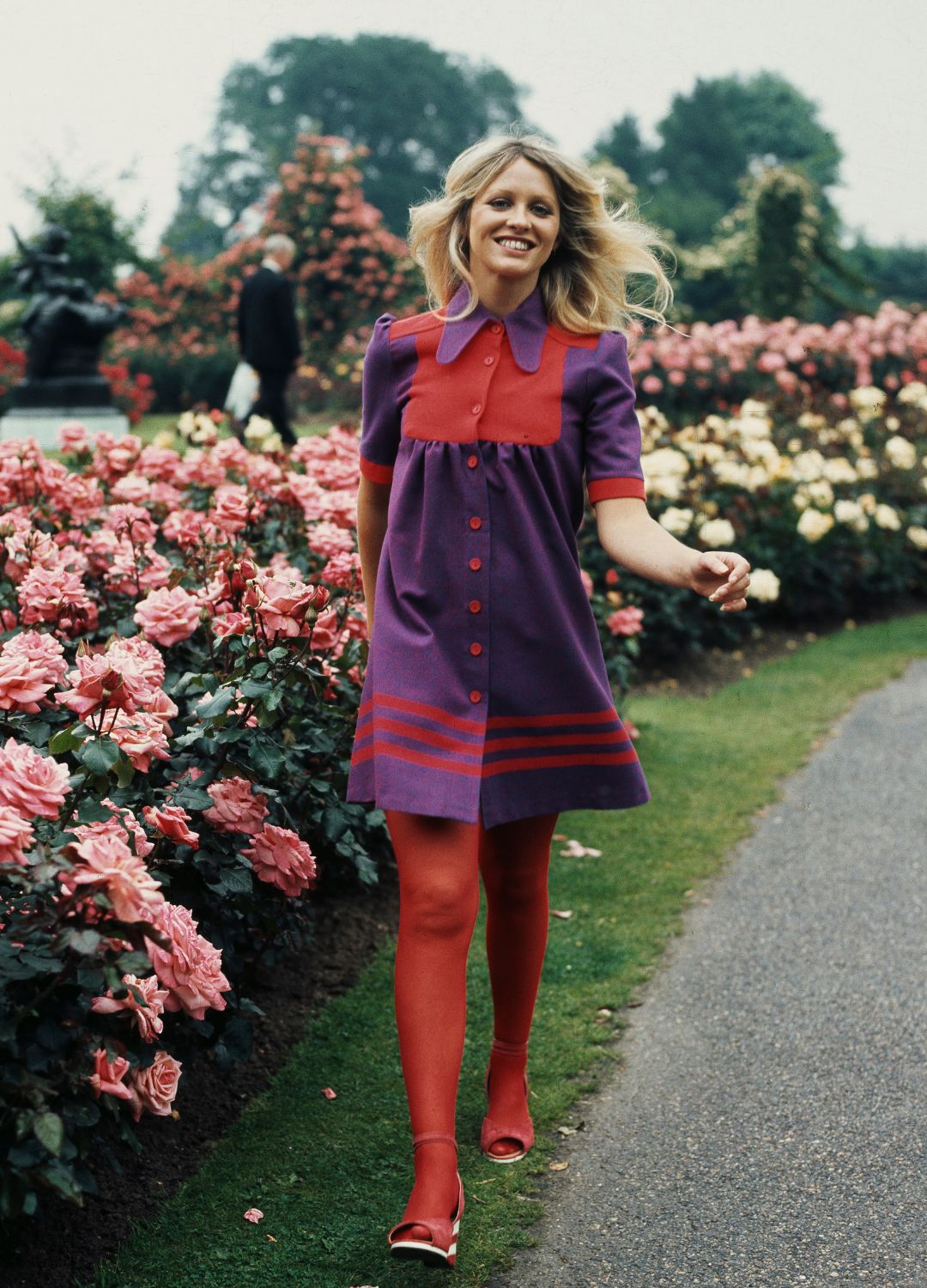A young woman wearing a red-and-purple dress with red stockings smiles as she walks beside rosebushes of pink roses, United Kingdom, circa 1975.