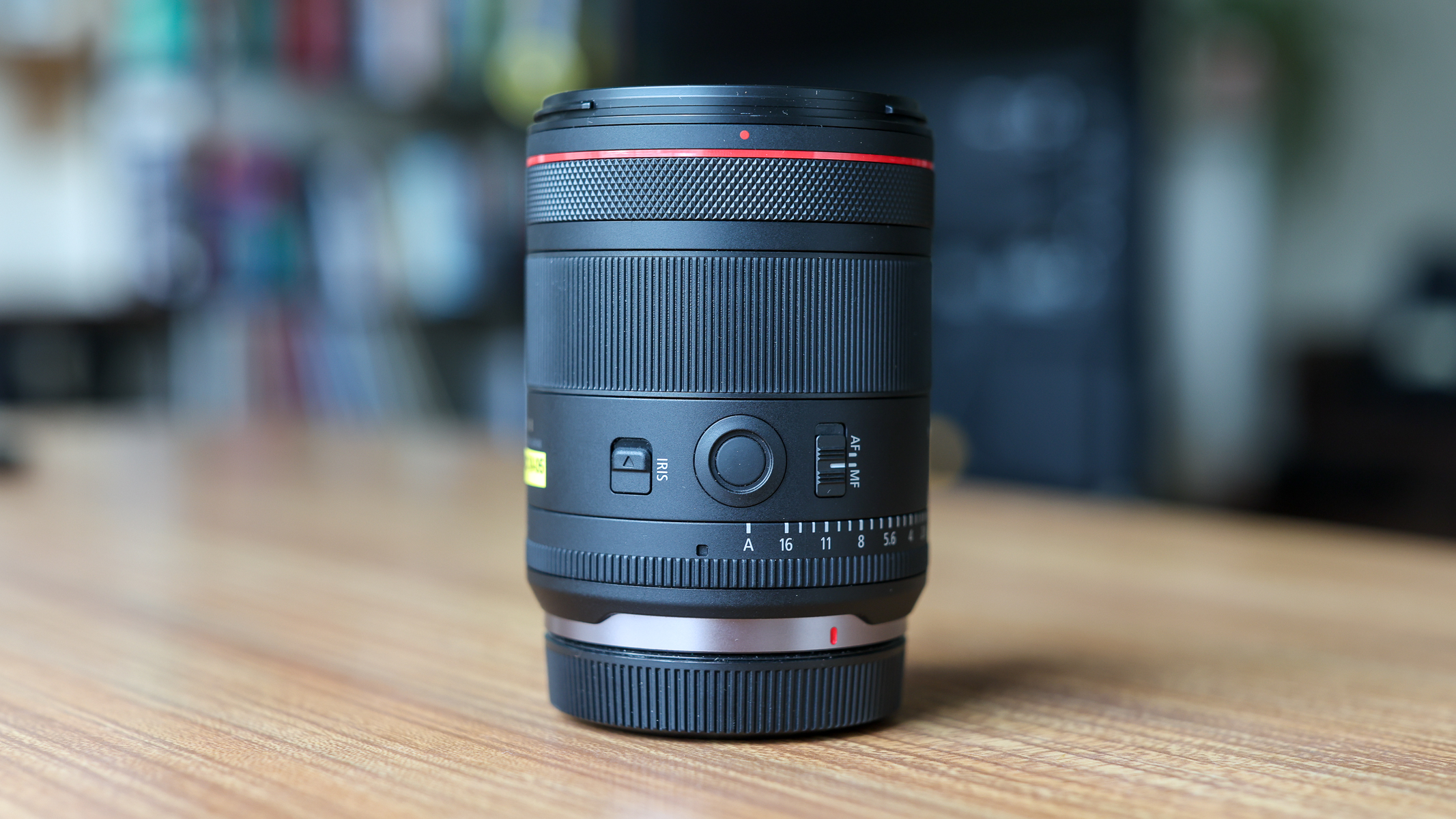 A side view of the Canon RF 20mm f/1.4 L VCM lens on a wooden table, showing aperture ring, iris lock and lens function button.
