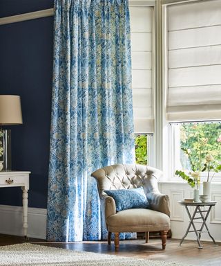 Blue living room with bay window, dressed with fabric blind and floral patterned curtains, brown armchair with cushion, dark wood flooring with rug, small gray wooden side table decorated with flowers