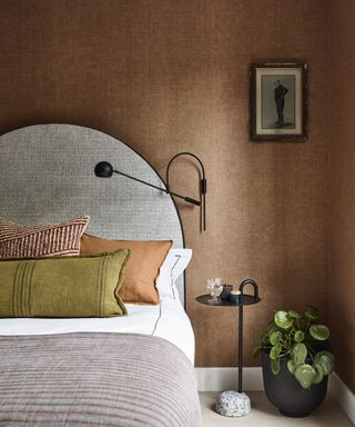 how to make a bedroom darker, earth bedroom style with textured ochre wallpaper, curved headboard, neutral bedding, green and ochre cushions, plant, modern black side table and wall light, artwork