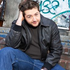 Photo of Michael Gandolfini sitting in front of a graffiti wall wearing a brown leather jacket with a dark sweater and blue jeans. He is running his hand through his hair.