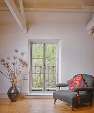 large white window in living space with comfy grey armchair, wooden floor and pewter vase on floor with twig and dried flower arrangement