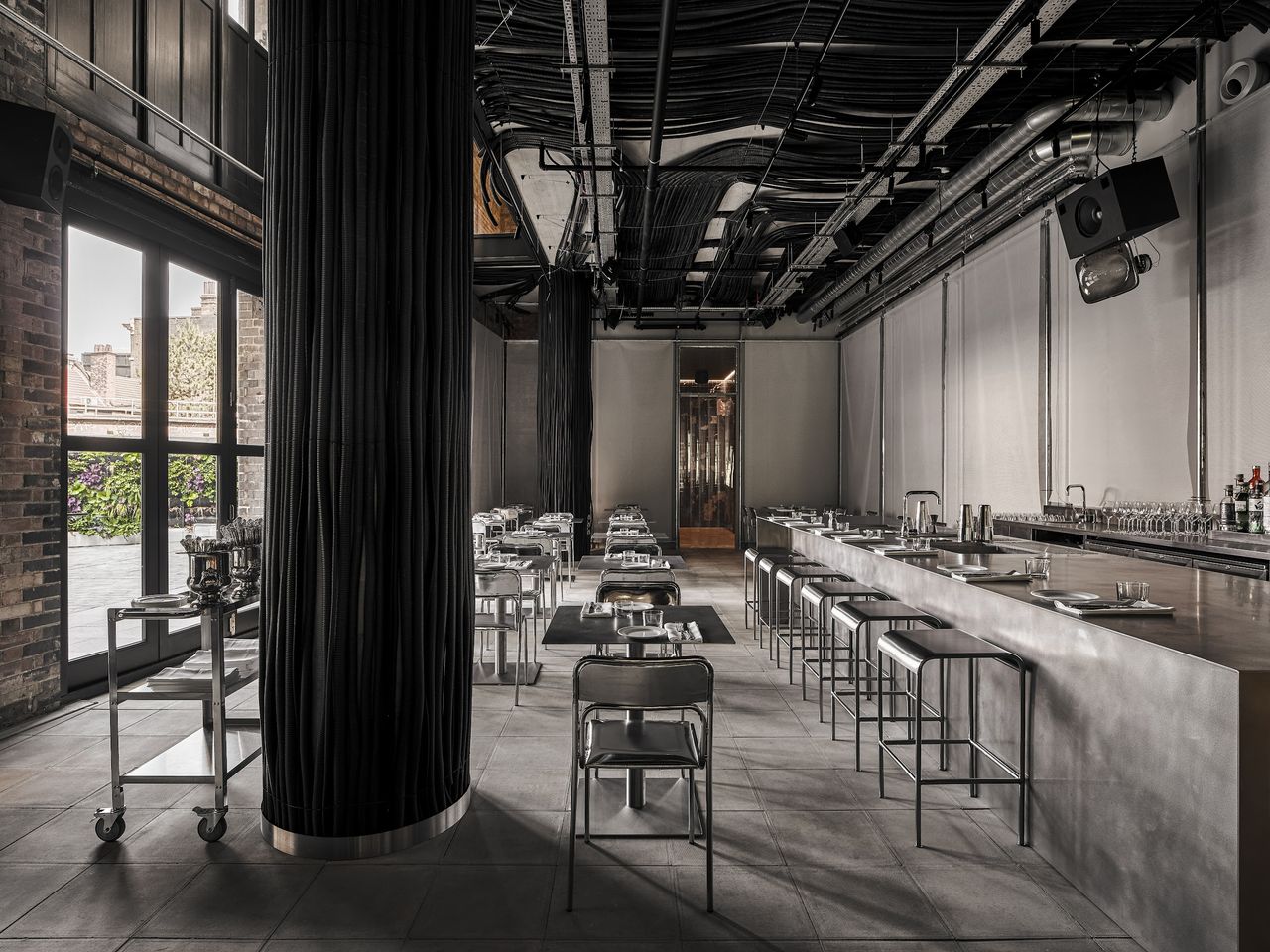 An industrial-chic restaurant features an exposed ceiling draped in wires, ventilation pipes, and cables, cement and brick walls, and chrome table, chair, and bar designs.