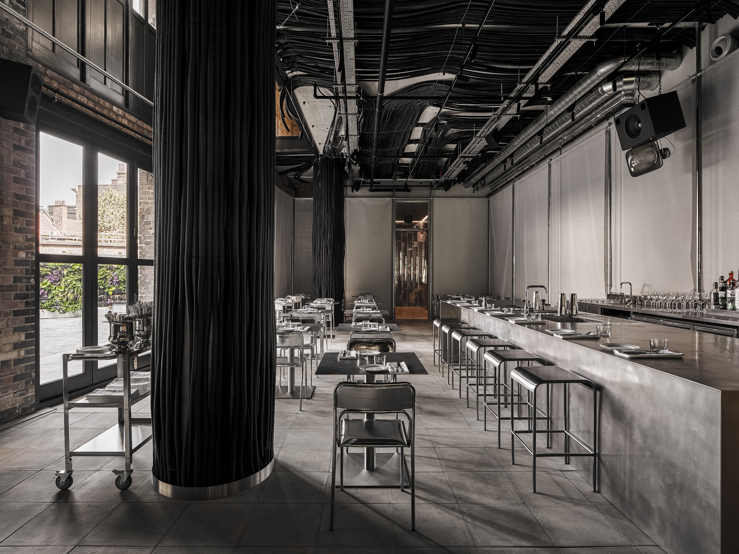 An industrial-chic restaurant features an exposed ceiling draped in wires, ventilation pipes, and cables, cement and brick walls, and chrome table, chair, and bar designs.