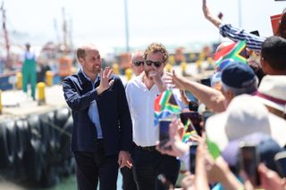 Prince William wearing a blue jacket and smiling waving to a huge crowd of people outside a harbor