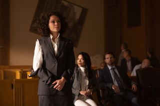 Jennifer Walters (Tatiana Maslany) in court, wearing a suit that has ripped on the shoulder on her right arm and is now barely attached, presumably the result of her Hulking out. A picture behind her on the wall is also now hanging at a strange 45-degree angle. Nonetheless, she is trying to present a composed, normal, lawyerly impression with her hands clasped in front of her.