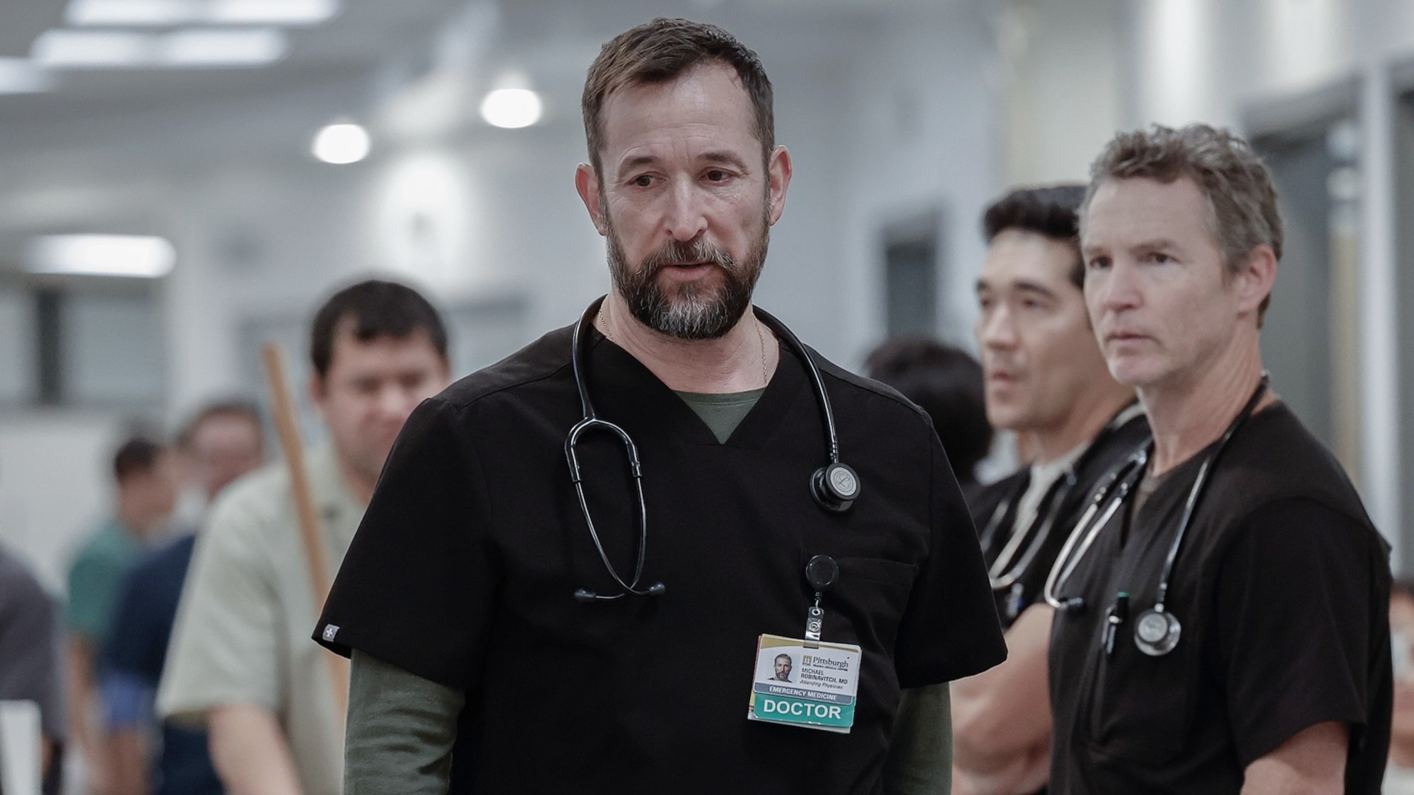 Noah Wyle walking in The Pitt with Shawn Hatosy and Ken Kirby standing behind him. 