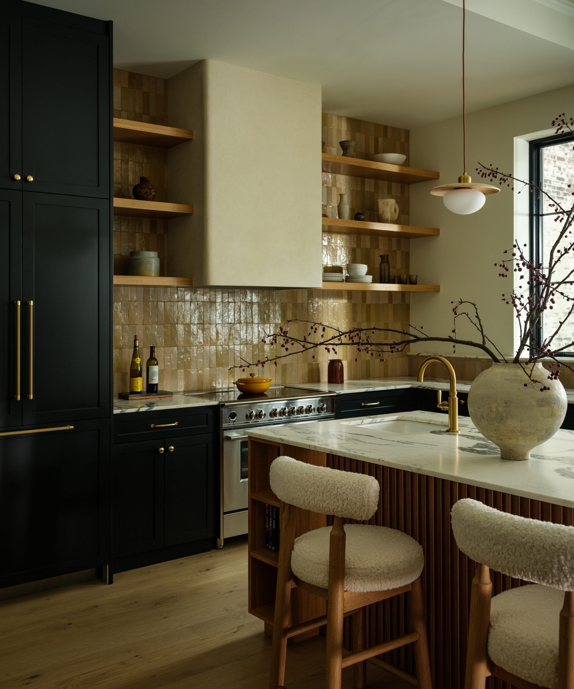 A kitchen with black cabinets, warm beige zellige backsplash tiles, a wooden island with boucle bar stools and marble countertops