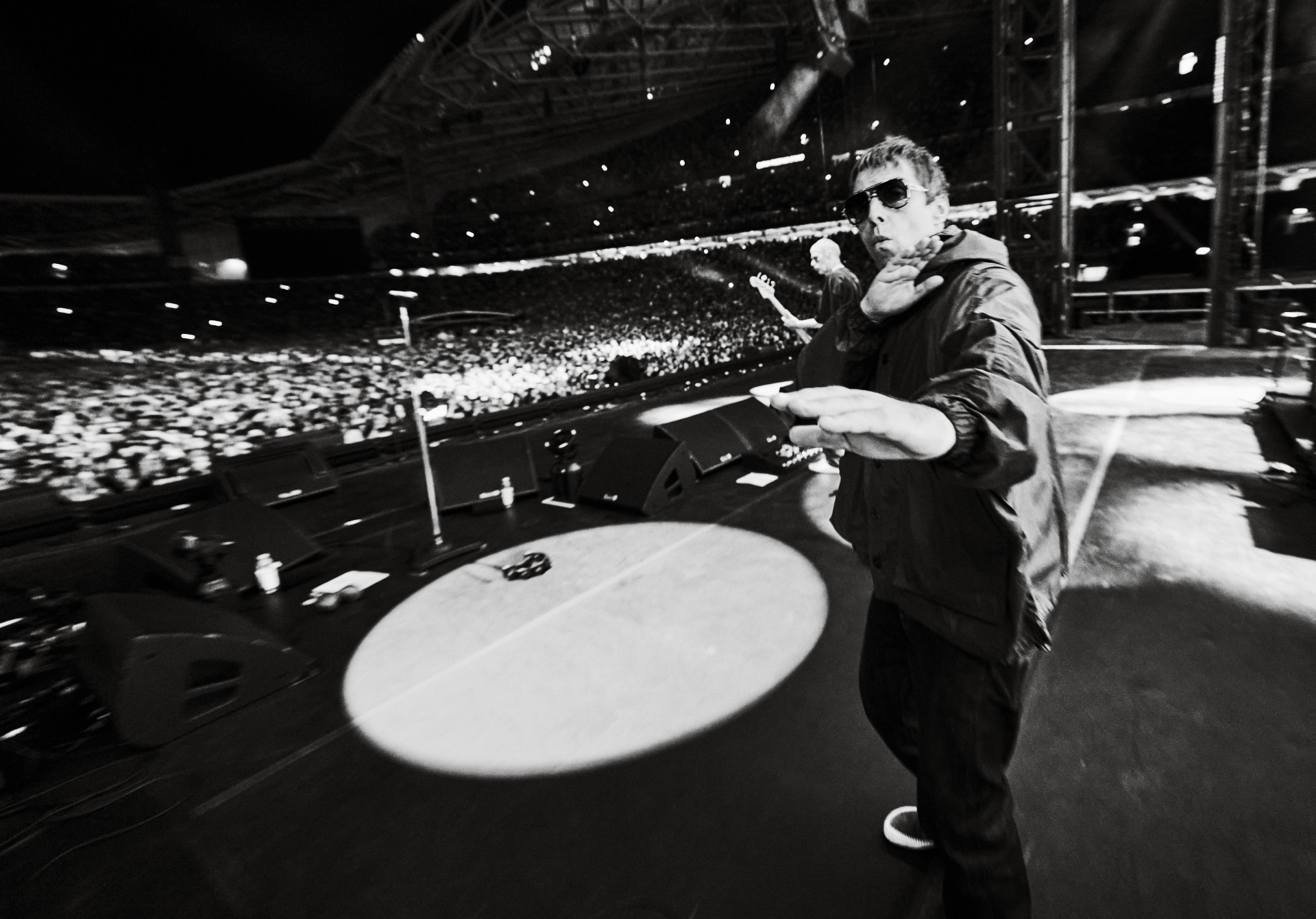 Liam Gallagher leans toward the camera with his arms outstretched during a performance in a massive, crowded stadium, captured in high-contrast black and white.