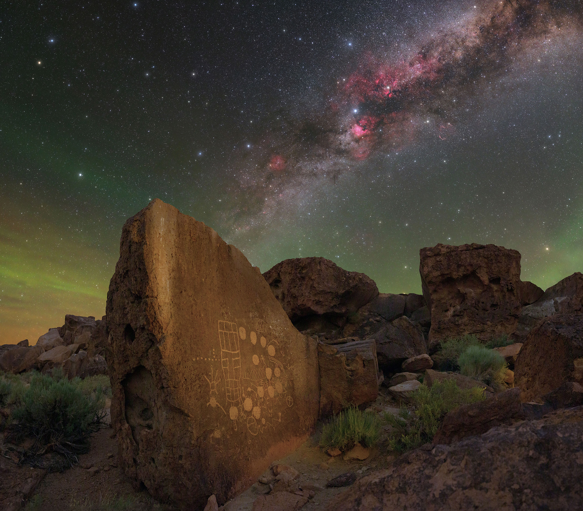 A pink galaxy is seen in the night sky over a series of rock formations.