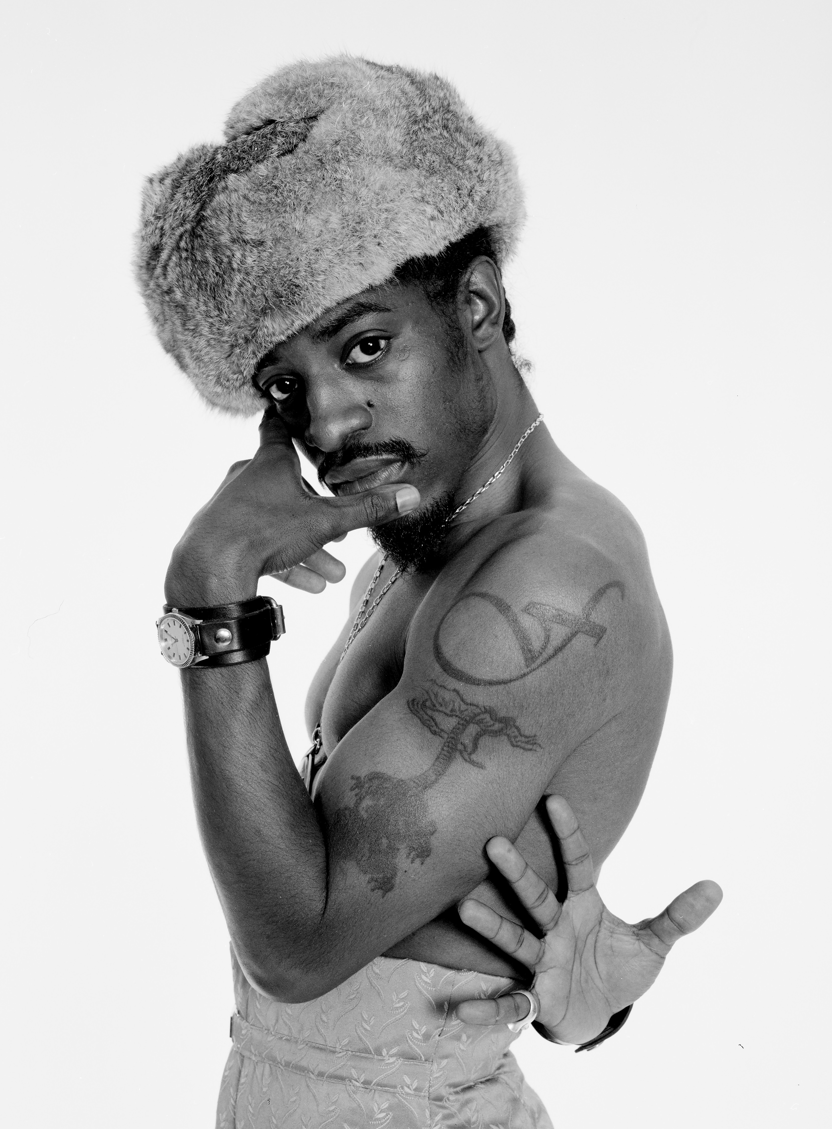 Black and white portrait of a shirtless Andre 3000 wearing a large fur hat, leaning on one arm and looking over his shoulder at the camera.