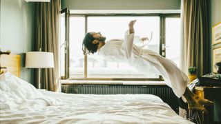 Man wearing a dressing gown jumps onto a bed covered with a white comforter