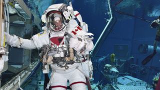 jeremy hansen in a spacesuit under water. he grasps on to a model of the international space station underwater. behind are divers and other space equipment