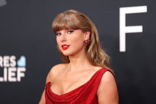 Taylor Swift wearing a red one shouldered dress and smiling in front of a gray background at Grammys 2025