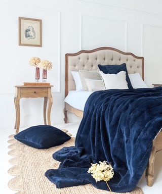 A bedroom with white paneled walls, a beige tufted bed with dark blue throw pillows and velvet throws, a wooden side table with two vases, and a jute scalloped runner rug
