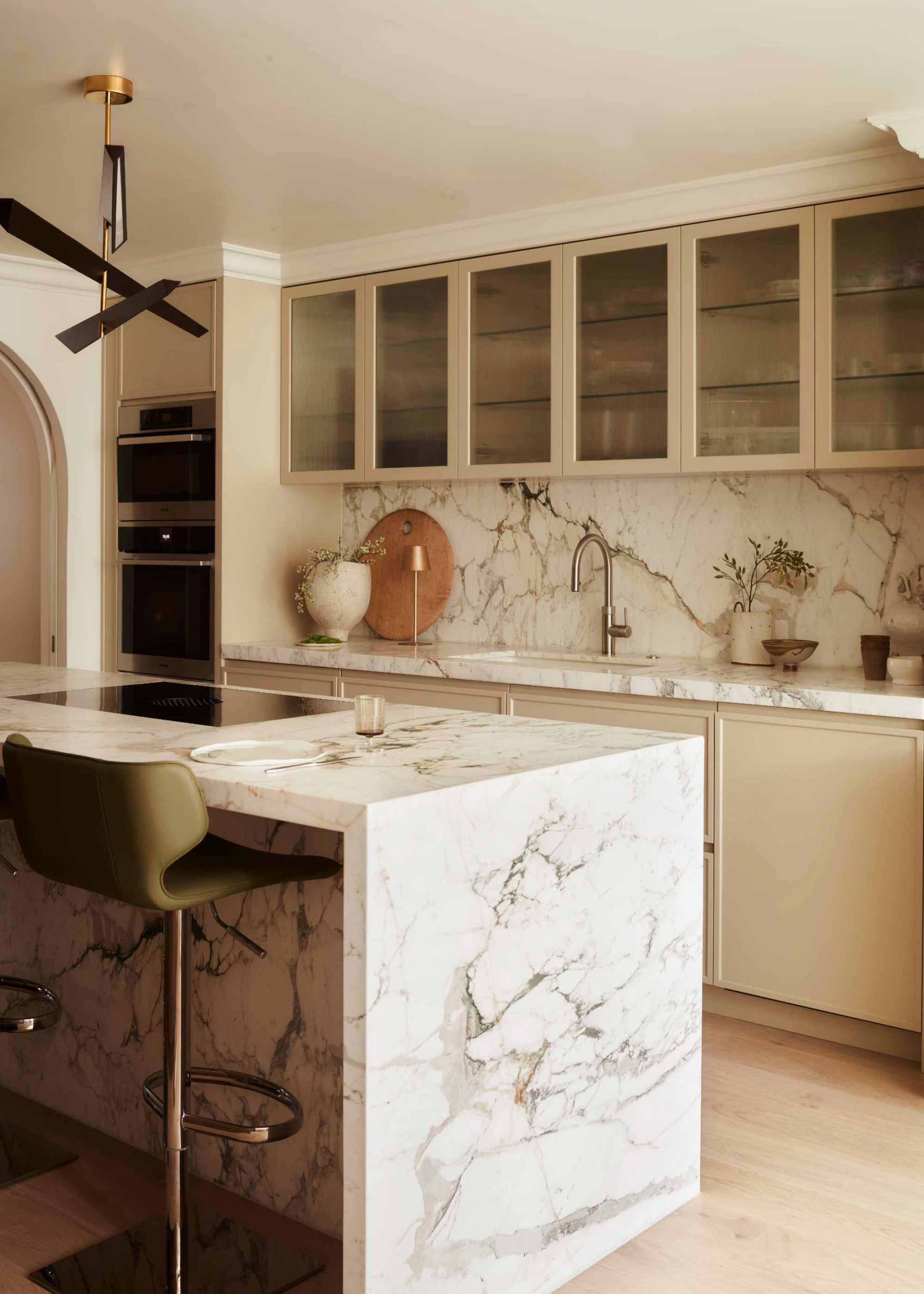 A minimalist kitchen with stone counters and a matching island, glass cabinets, and a green stool