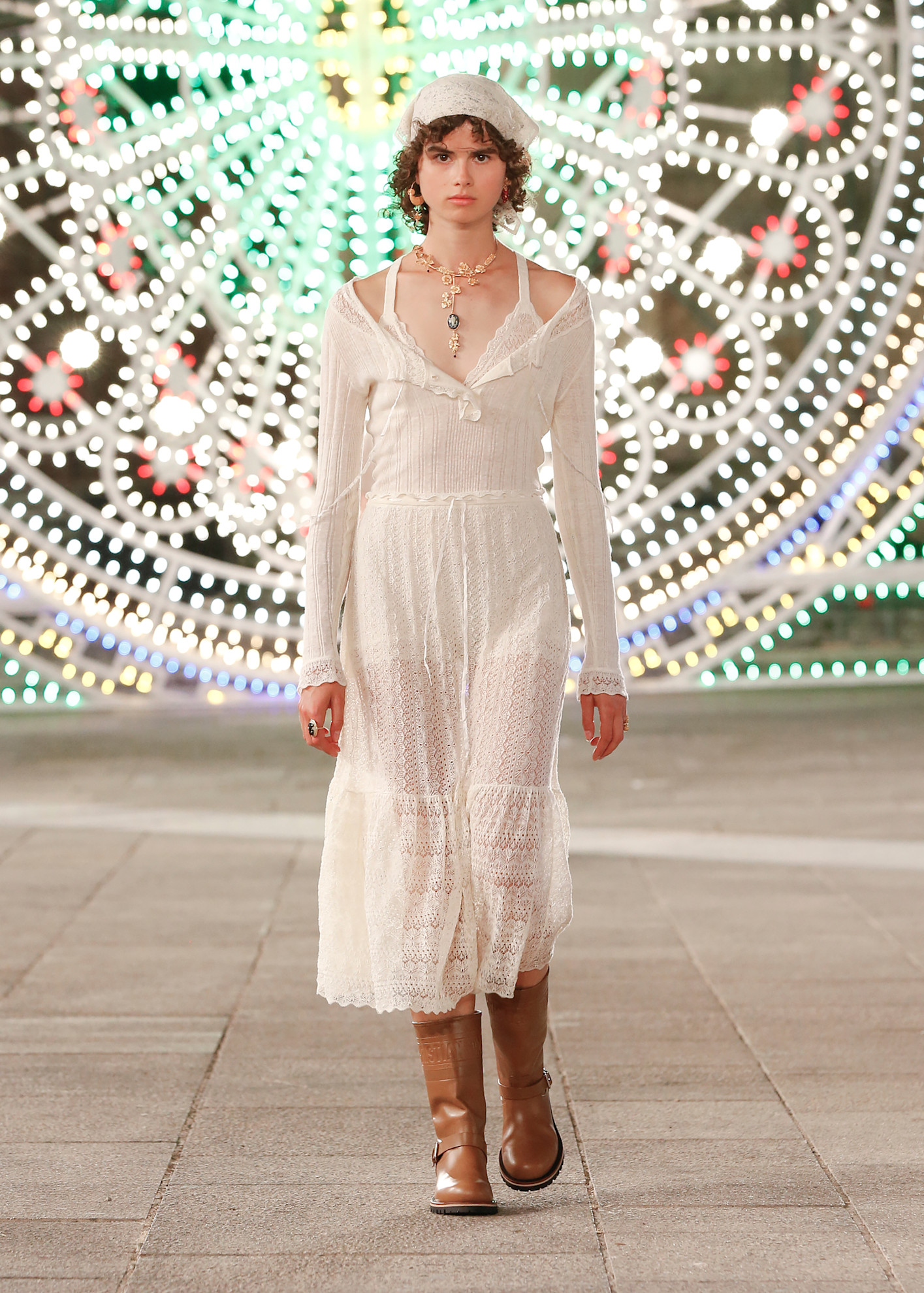 The Dior Autumn / Winter 2021 Cruise collection. The model wears a lace dress, slouch boots and a lace headscarf. This image is from Launchmetrics.