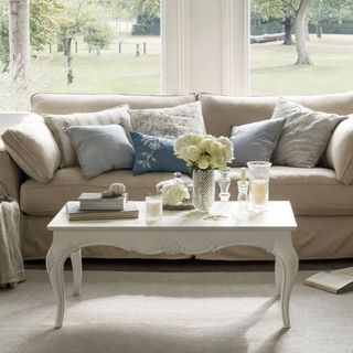 living room with cream colour sofa and designed cushion glass window and flower vase on white table