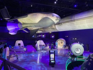 A full-scale model of Sierra Space's Dream Chaser spaceplane with "Shooting Star" cargo module is suspended above other exhibits in "Gateway: The Deep Space Launch Complex."