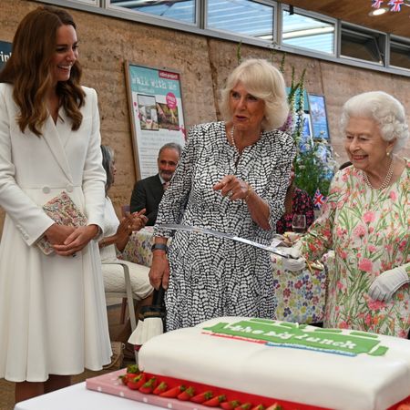 Princess Kate wears a white coat dress and has long brown hair, she laughs as Queen Camilla, wearing a black and white patterned dress, points and Queen Elizabeth holds a large knife
