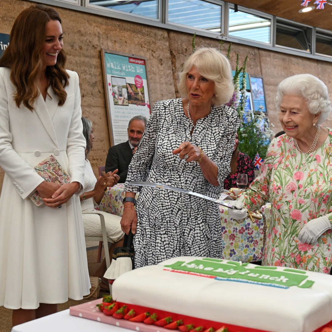 Princess Kate wears a white coat dress and has long brown hair, she laughs as Queen Camilla, wearing a black and white patterned dress, points and Queen Elizabeth holds a large knife