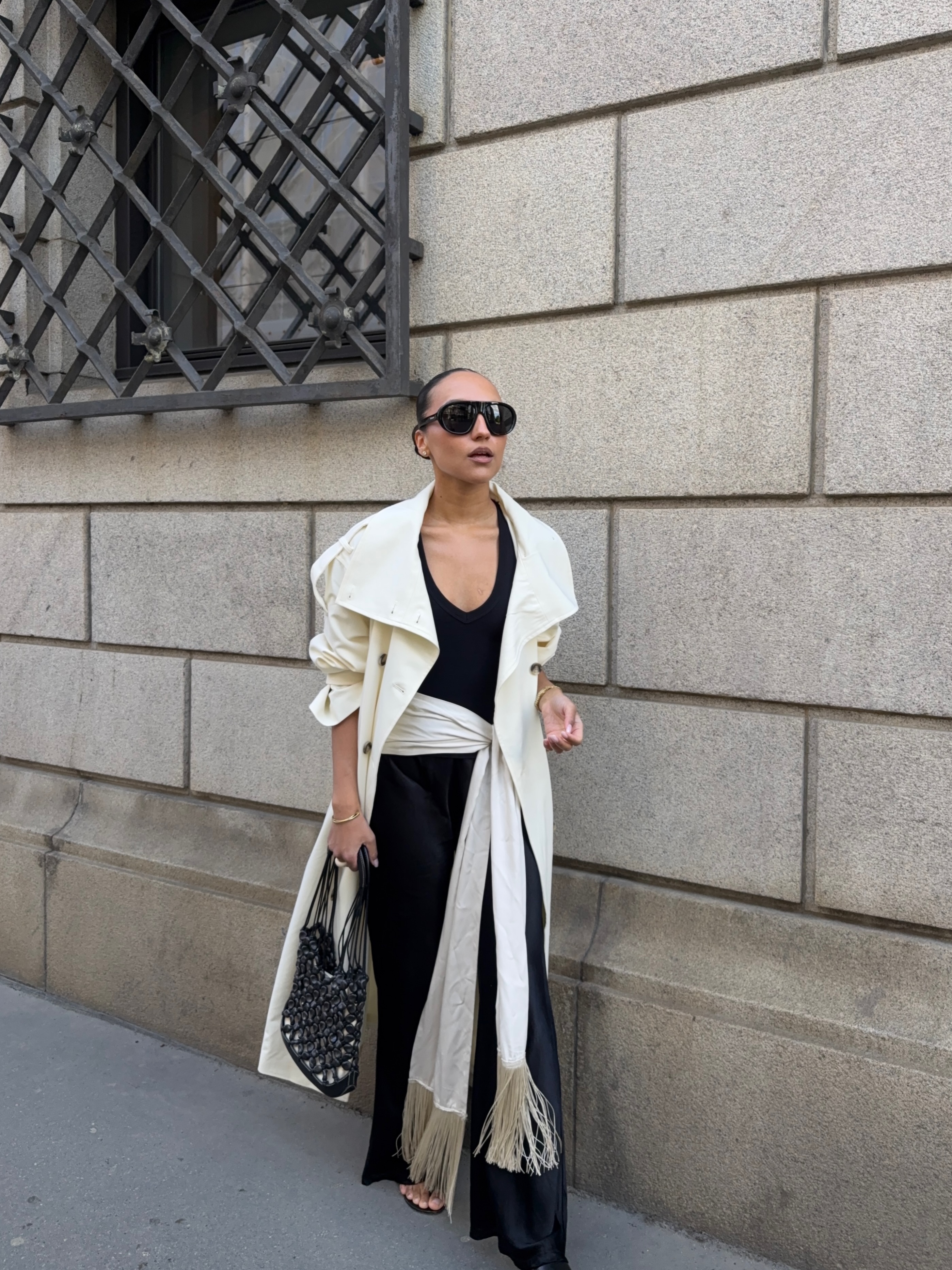 An image of an influencer wearing a cream long trench coat over a black top, black satin trousers and a satin tassel scarf, carrying a black woven handbag.