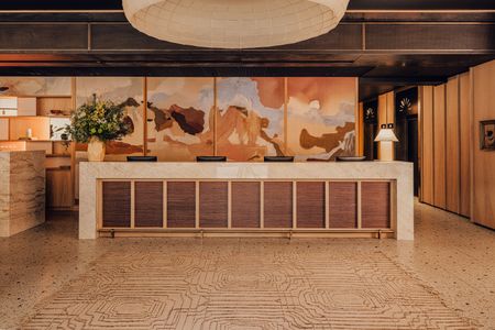 A warmly lit hotel lobby with a central marble and wood counter decorated with a vase, on the left, and a lamp, on the right, in similar hues, a handpainted mural behind it, speckled terrazzo flooring, and textural surfaces throughout.