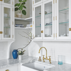 Corner detail of white fitted kitchen with glass cupboard doors and brass fittings including vintage taps over Belfast sink surrounded by marble work top.