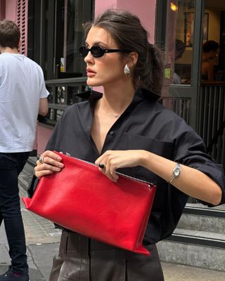 British fashion influencer Valera @sobalera poses on a sidewalk in London wearing black oval sunglasses, silver tear drop earrings, a black button-down shirt, vintage wavy watch, and red clutch bag