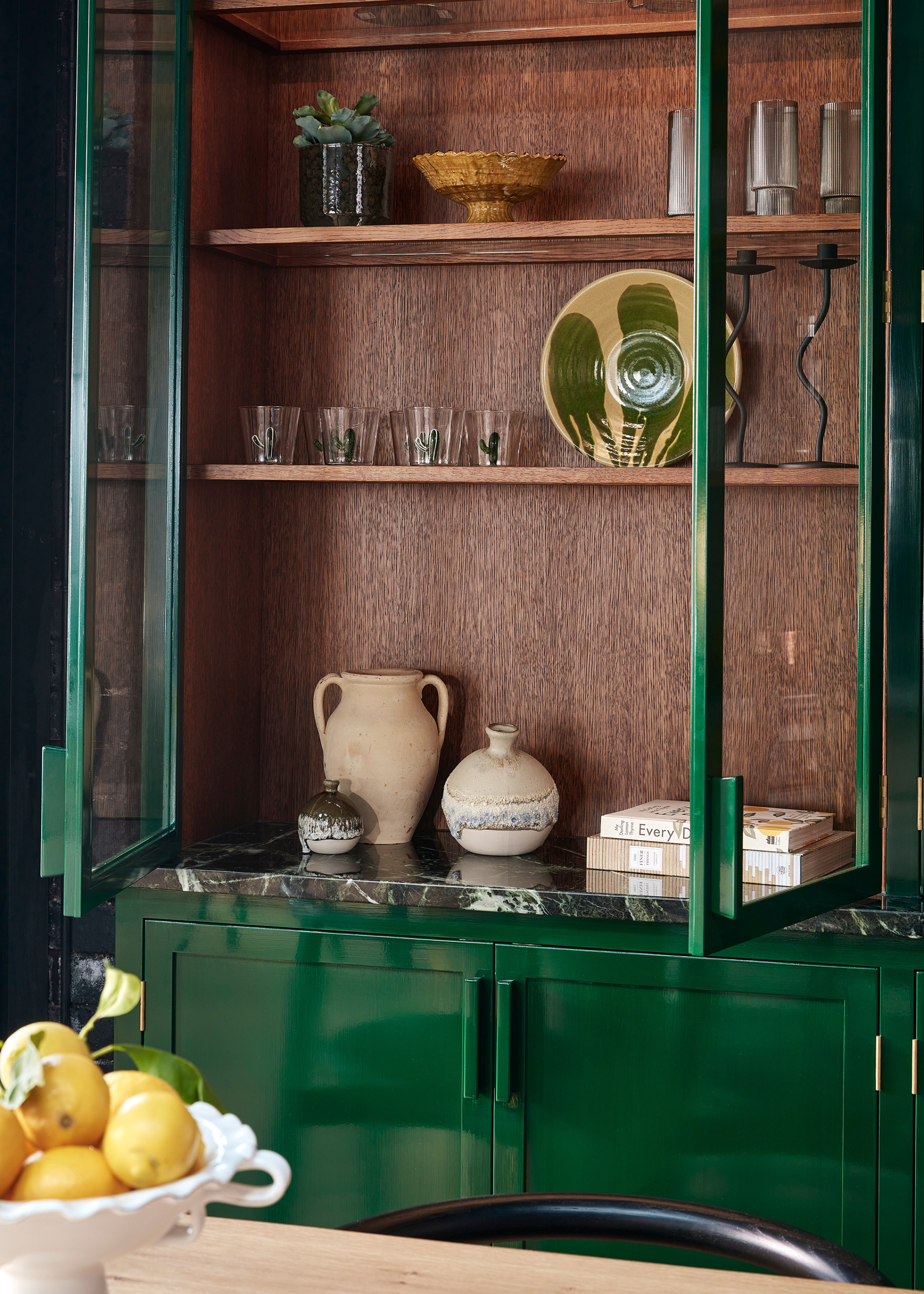 Green gloss, glass-fronted kitchen cabinets that are open and filled with decorative glassware and tableware in front of a natural wood table with a fruit bowl on top