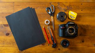 A top-down shot of black card, scissors, Nikon DSLR, tape measure, scalpel, pencil, sticky tape, 50mm lens, and DIY lens hood on wooden surface