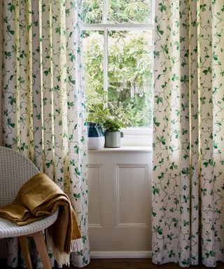 Large window looking out to green garden dressed with white and green leaf patterned curtains, chair with yellow throw