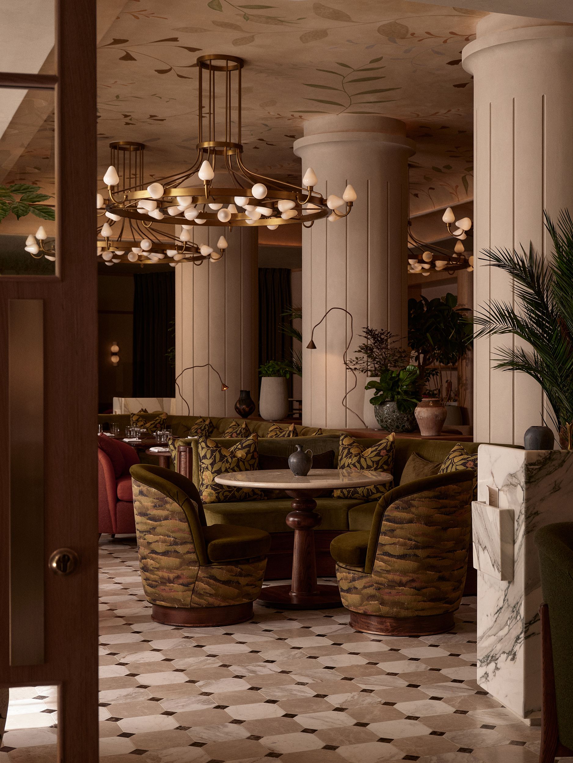 A beautifully decorated hotel bar features slanted wood and marble tables, upholstered armchairs with a camouflaged fabric and a cocooning structure, huge columns, and chandeliers with bulb-shaped light bulbs.