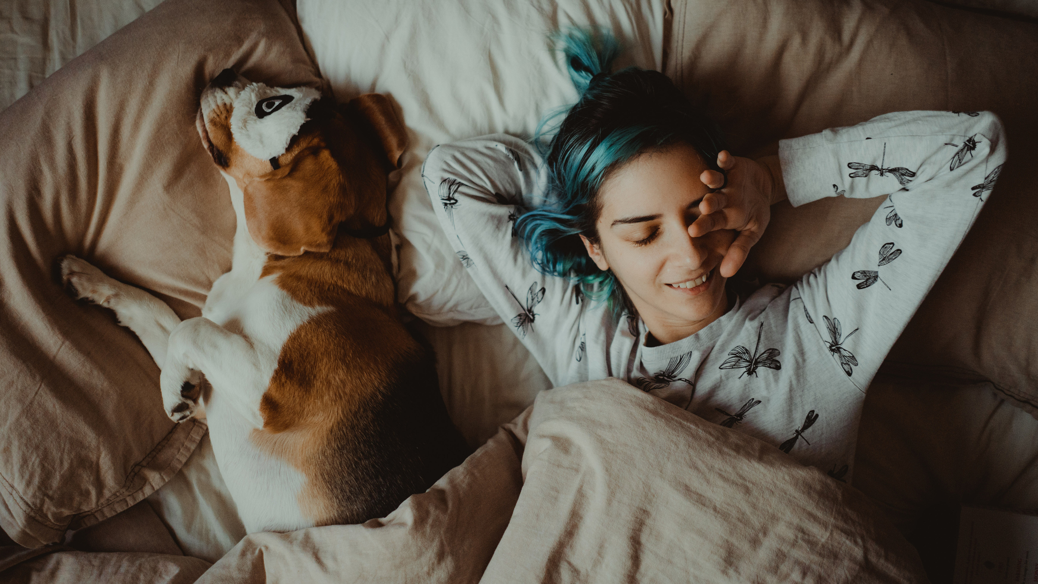 A woman lies in bed next to her dog that's wearing a patterned eye mask