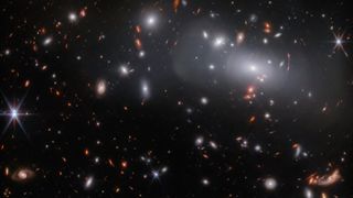 A large elliptical galaxy surrounded by many smaller similar galaxies as seen by the James Webb Space Telescope.